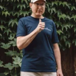 Mann mit Cap und Brille zeigt Daumen hoch vor grüner Efeuwand.