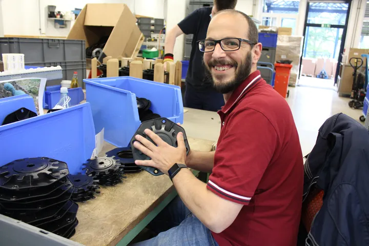 Lächelnder Mitarbeiter montiert Bauteile an Werkbank mit Sortierboxen in der Werkstatt.