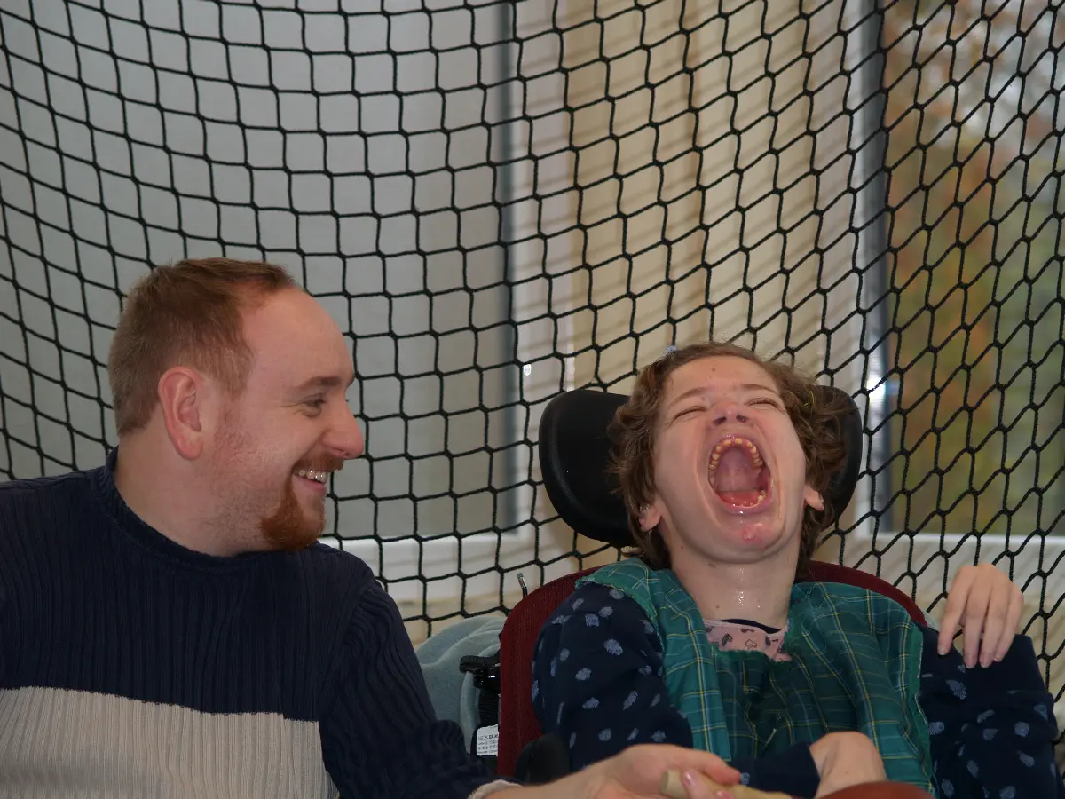 Mann lacht mit fröhlichem Jugendlichen im Rollstuhl in inklusiver Freizeitsituation.