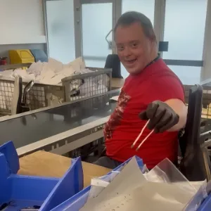Lächelnder Mitarbeiter sortiert Verpackungsmaterial an Montage- und Verpackungstisch in Produktionshalle.