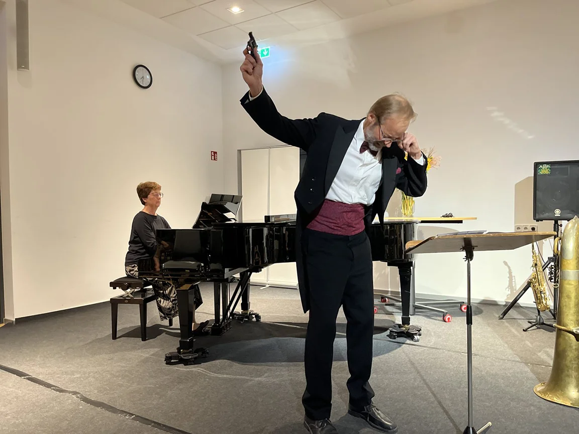 Solist im Frack mit Pistolenrequisite, begleitet von Pianistin bei Klavierabend im Konzertsaal.