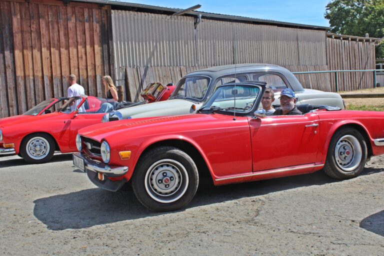 Roter Triumph TR6 Cabrio bei Oldtimer-Ausfahrt, Fahrer und Beifahrer lächeln in der Sonne.