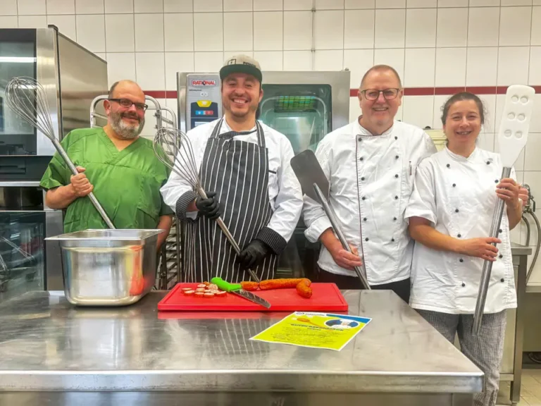 Küchenteam in Großküche lächelt mit Kochutensilien an Edelstahltheke.