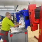 Mitarbeiter sortiert Papier, während Kippgerät Container in Recyclinghalle entleert.