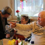 Lachende Senioren im Pflegeheim am Tisch mit Pflegerin und Frühlingsblumen.
