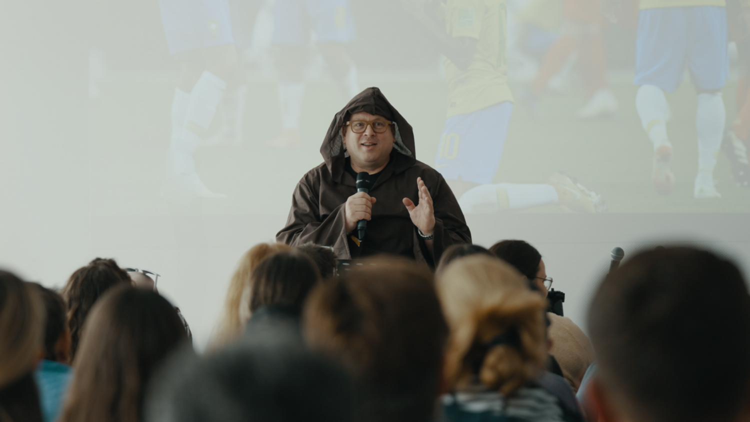 Pastor predigt mit Mikrofon vor Publikum, Fußballspiel als Folie im Hintergrund