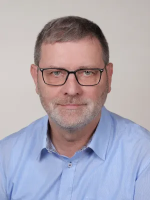 Professionelles Porträt eines Mannes mit Brille, grauem Bart und blauem Hemd.