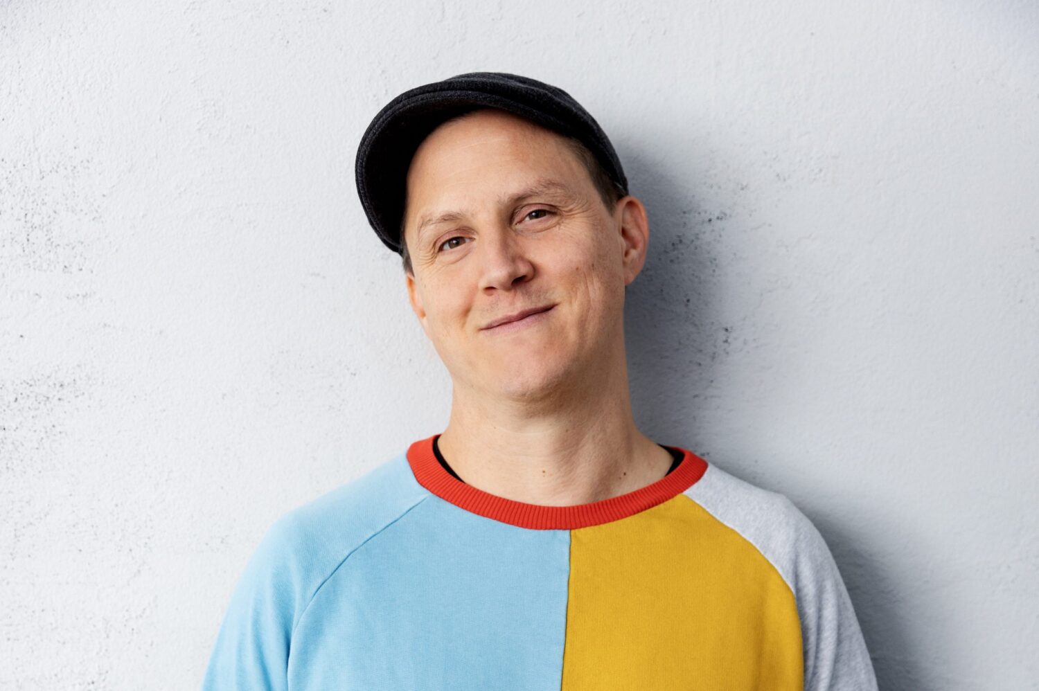 Porträt von Sebastian mit Schiebermütze und Farbblock-Pullover vor grauer Wand.