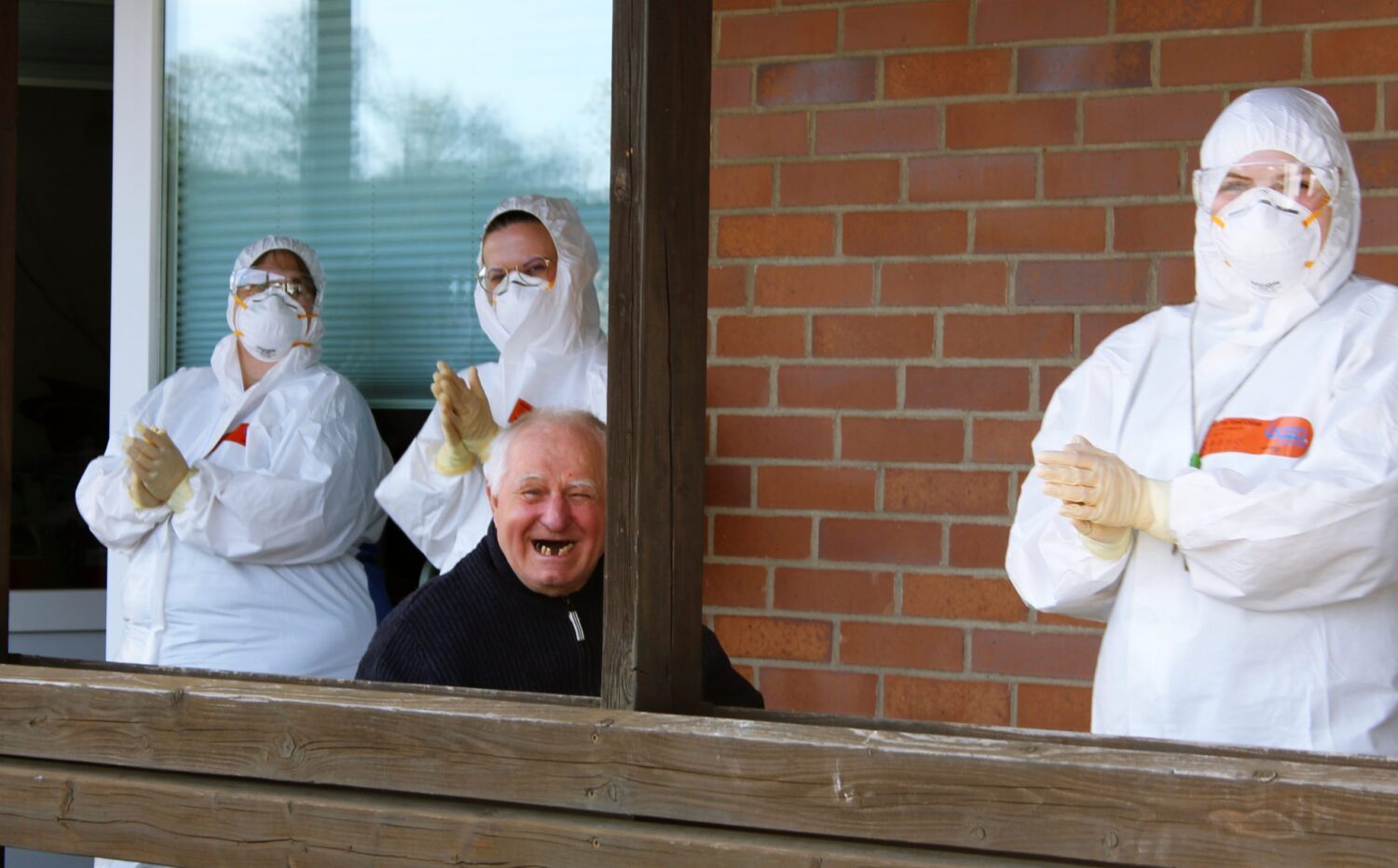 Senior lächelt glücklich, umgeben von Pflegekräften in Schutzkleidung vor dem Wohnheim.