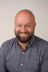 Bärtiger, glatzköpfiger Mann lächelt freundlich im Business-Porträt vor hellem Hintergrund.