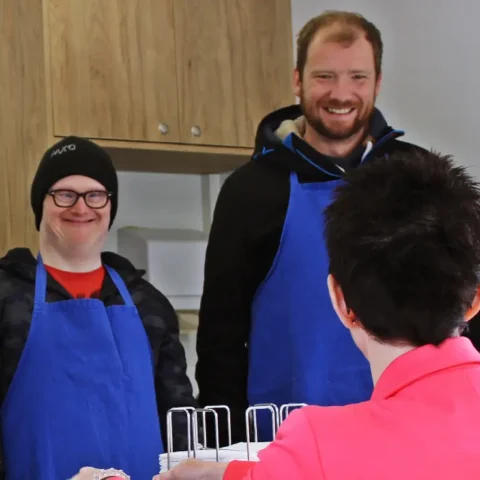 Zwei Männer im blauen Schürzen-Team bedienen Kundin am Suppenstand in der Küche.