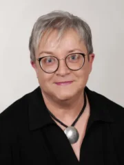 Porträt von Tanja Rockensüß mit Brille, grauen Haaren und modernem Anhänger vor neutralem Hintergrund.