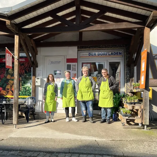 Team in grünen Schürzen vor dem Dorfladen mit Café, regionale Lebensmittel und Pflanzen.