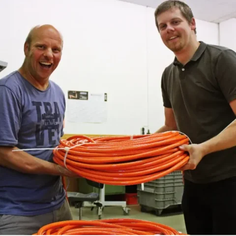 Zwei Techniker halten lächelnd eine große orange Kabelrolle in Werkstatt.