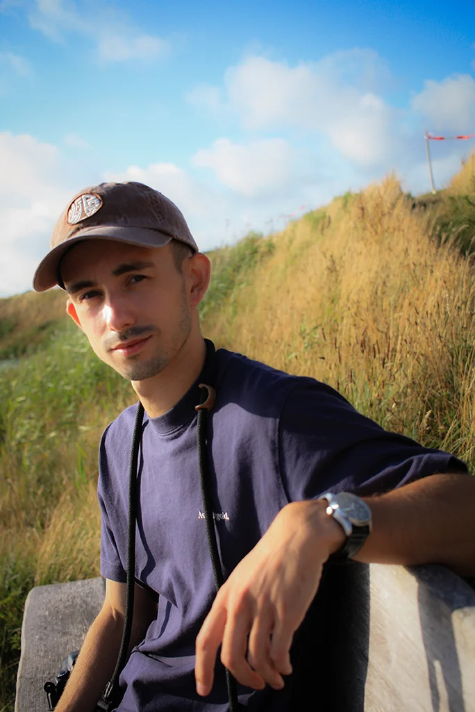 Junger Mann mit Kamera und Baseballcap sitzt in goldenem Abendlicht in Graslandschaft.
