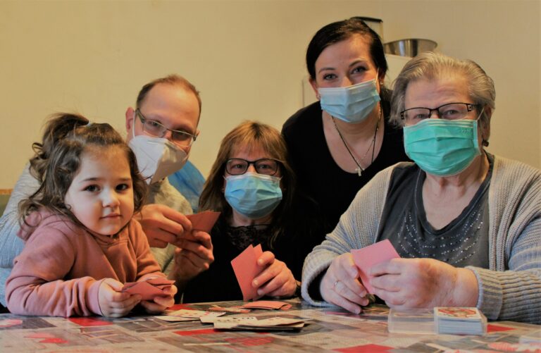 Mehrgenerationen-Familie spielt Kartenspiel mit Masken am Küchentisch, Mädchen schaut in die Kamera.