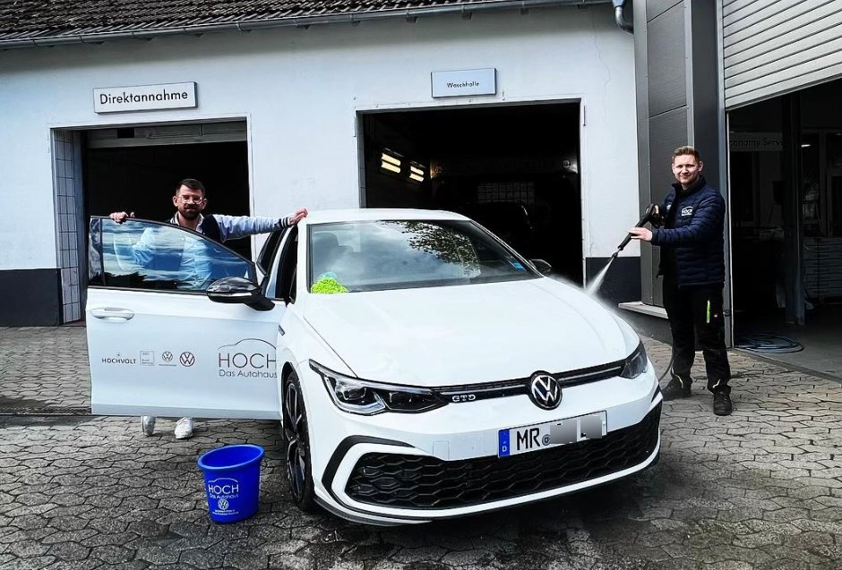 Zwei Mitarbeiter reinigen einen weißen VW GTD mit Hochdruckreiniger vor Autohaus Hoch.