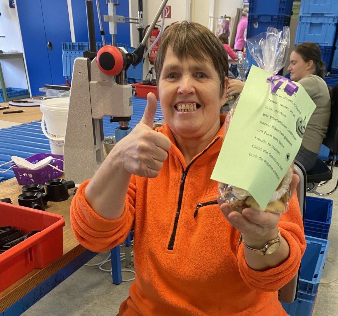 Werkstattmitarbeiterin zeigt Daumen hoch und hält Keks-Geschenk mit Dankeskarte.