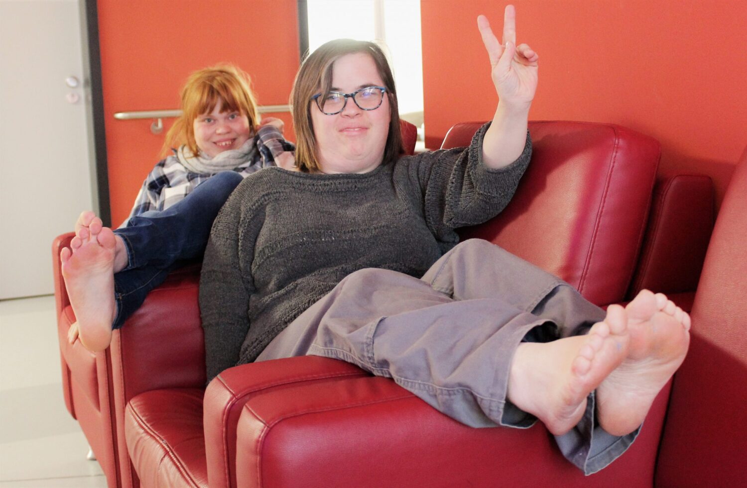 Zwei Frauen entspannen barfuß auf roten Sesseln vor orangefarbener Wand und lächeln in die Kamera.