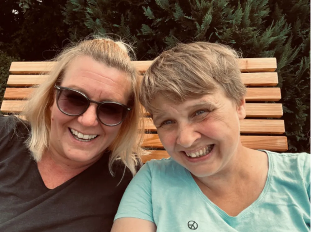 Zwei Freundinnen lächeln auf Holzbank im Grünen, eine mit Sonnenbrille.