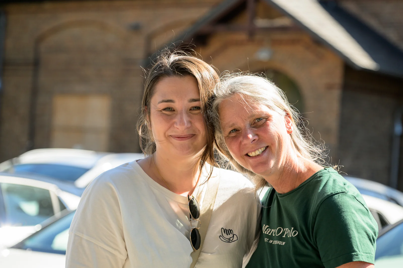 Zwei Frauen lächeln im Sonnenschein vor Backsteingebäude, urbaner Hintergrund mit Autos.