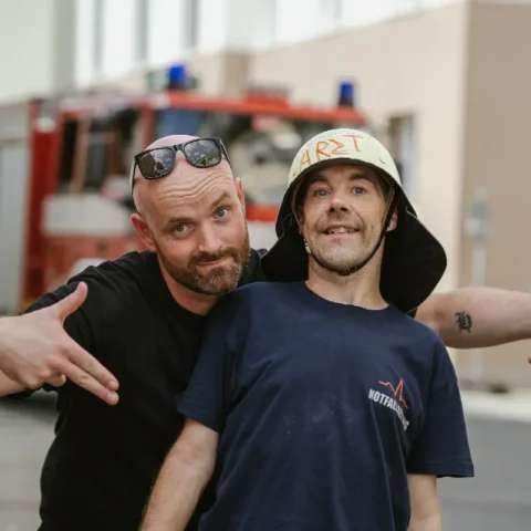 Zwei Männer lachen vor Feuerwehrauto, einer trägt Feuerwehrhelm mit Aufschrift Arzt.