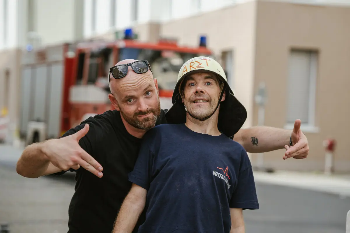 Zwei Männer lachen vor Feuerwehrauto, einer trägt Feuerwehrhelm mit Aufschrift Arzt.